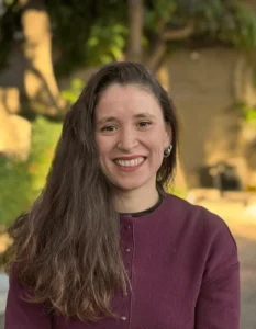 Retrato de Daniela Dinamarca Figueroa sonriendo, con cabello largo oscuro y vistiendo un cárdigan color burdeo en un entorno al aire libre.