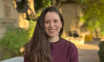 Retrato de Daniela Dinamarca Figueroa sonriendo, con cabello largo oscuro y vistiendo un cárdigan color burdeo en un entorno al aire libre.