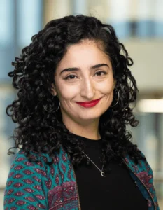 Retrato de María José Lagos Serrano sonriendo, con cabello rizado y oscuro, vistiendo una chaqueta estampada turquesa en el interior de un edificio moderno.