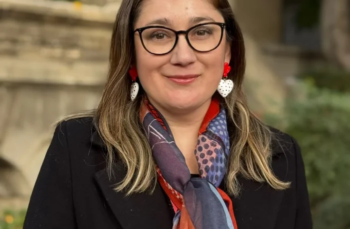 Retrato de Daniela Avello con gafas, chaqueta negra y bufanda colorida en exteriores.