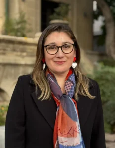 Retrato de Daniela Avello con gafas, chaqueta negra y bufanda colorida en exteriores.