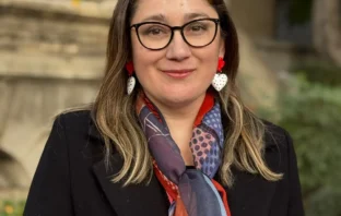 Retrato de Daniela Avello con gafas, chaqueta negra y bufanda colorida en exteriores.