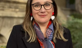 Retrato de Daniela Avello con gafas, chaqueta negra y bufanda colorida en exteriores.