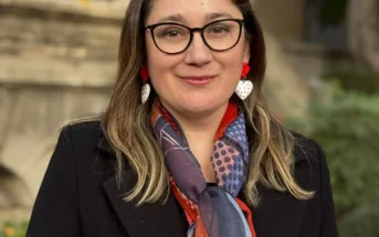 Retrato de Daniela Avello con gafas, chaqueta negra y bufanda colorida en exteriores.