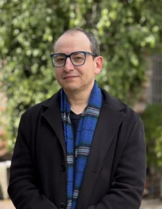 Retrato de académico Sergio Galdames con gafas, chaqueta negra y bufanda colorida en exteriores.